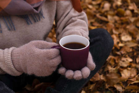 Gloved hands hold a mug of tea, against the backdrop of autumn foliage. Late autumn.の写真素材