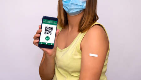a girl in a medical mask shows QR code and vaccination certificate on her phoneの写真素材
