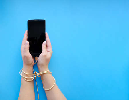 A close-up view from above of womens hands connected by a wired cable with a smartphone gadget, on a blue background.の写真素材