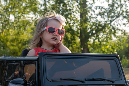 a little girl is riding a childrens car in the park, talking on the phone.