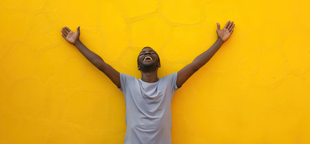 Happy african american young man with hands up against yellow wallの素材