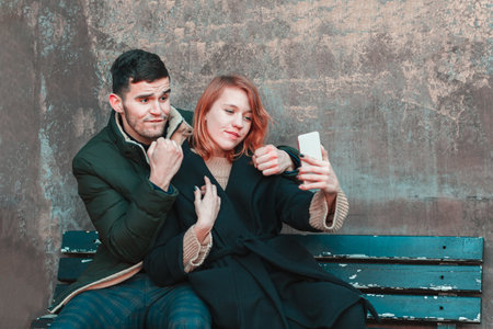 Cheerful Emotional Young Couple Sitting on the Bench and Making Selfie. Two Happy People Love Story on the Streetの写真素材