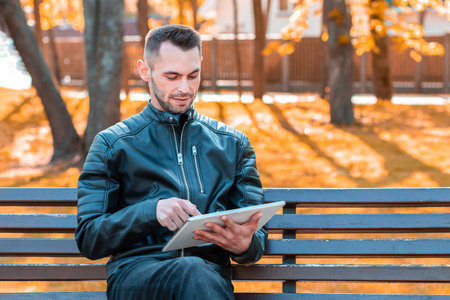 Handsome Young Man Sitting on the Bench and Using Big White Tablet PC at the Beautiful Autumn Parkの写真素材