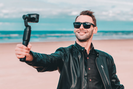 Portrait of Smiling Blogger in Sunglasses Making Selfie or Streaming Video at the Beach Using Action Camera with Gimbal Camera Stabilizer. Man in Black Clothes Making Photosの写真素材