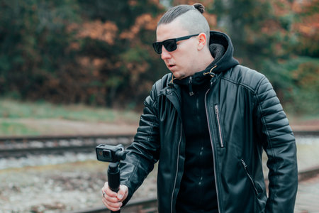 Youthful Blogger in Black Clothes and Sunglasses Making Video Using Action Camera with Gimbal Camera Stabilizer in Pine Forestの写真素材