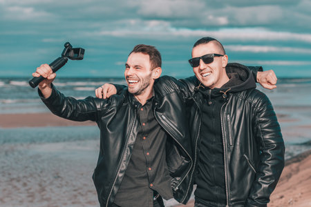 Two Handsome Smiling Friends Making Selfie Using Action Camera with Gimbal Stabilizer at the Beach. Youthful Men in Black Clothes Having Fun by Making Photosの写真素材