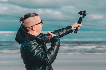 Young Blogger in Sunglasses Making Selfie or Streaming Video at the Beach Using Action Camera with Gimbal Camera Stabilizer. Hipster in Black Clothes Making Photo Against the Seaの写真素材