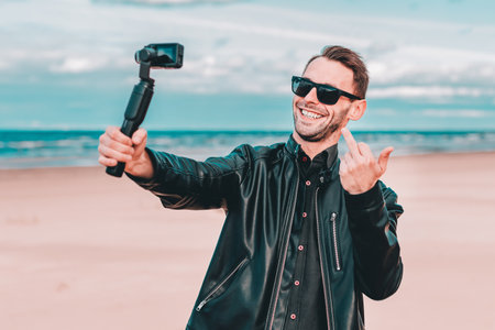 Youthful Blogger in Sunglasses Making Selfie or Streaming Video at the Beach Using Action Camera with Gimbal Camera Stabilizer. Handsome Man in Black Clothes Making Photo Against the Seaの写真素材