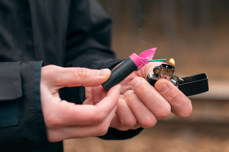 Setting Fire to the Firecracker. Man in Black Clothes Lighting Up the Petard. Firing Up the Pyrotechnics with a Black Gasoline Lighter Outdoorsの写真素材