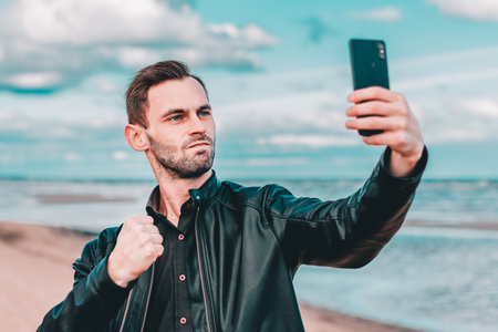 Young Blogger Making Selfie or Streaming Video at the Beach Using Black Smartphone. Handsome Man in Black Clothes Making Photo Against the Sea at Cloudy Dayの写真素材