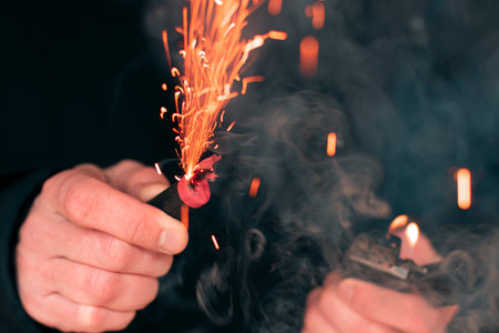 The Firecracker in a Hand. Man Holding a Burning Petard in His Hand. A Human with a Pyrotechnics that Burns with Sparks and Smoke Outdoorsの写真素材