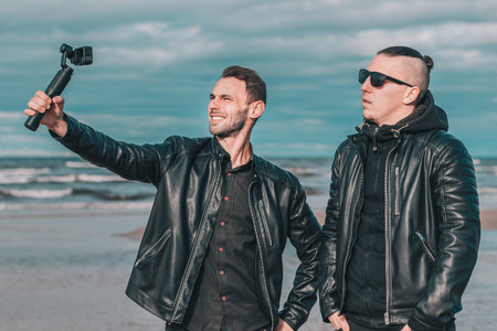 Two Handsome Smiling Friends Making Selfie Using Action Camera with Gimbal Stabilizer at the Beach. Youthful Men in Black Clothes Having Fun by Making Photosの写真素材
