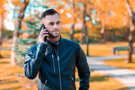 Handsome Young Man Talking on the Phone at the Beautiful Autumn Park. Youthful Guy Using Smartphone for the Call Outdoors at Sunny Dayの写真素材
