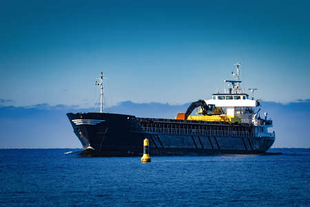 Black Sailing Bulk Carrier. Cargo Ship with Long Reach Excavator Moving in Still Water at Sunny Day by the Seaの写真素材