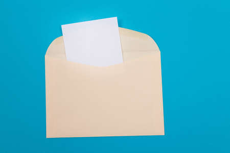 Beige Envelope with Blank White Sheet of Paper Inside, Lying on Blue Background - Mock Up with Copy Space. Receiving Mail or Notice, Sending Postcard - Top View, Flat Layの写真素材