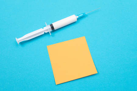 Vaccination, Immunology or Revaccination Concept - A Medical Disposable Syringe Lying on Blue Table in Doctors Office in a Hospital or Clinic. Blank Orange Sticky Note - Mock Up with Copy Spaceの写真素材