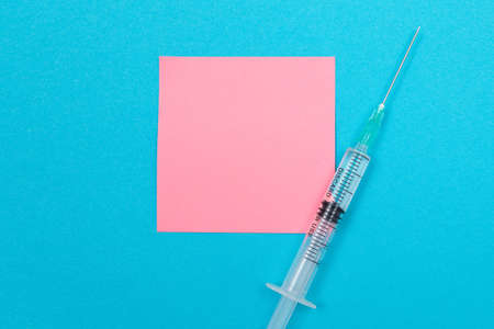 Vaccination, Immunology or Revaccination Concept - A Medical Disposable Syringe Lying on Blue Table in Doctors Office in a Hospital or Clinic. Blank Pink Sticky Note - Mock Up with Copy Spaceの写真素材