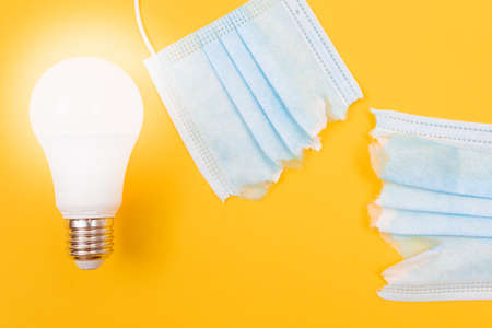 Glowing Power Saving Lamp with Torn Medical Face Mask Lying on Yellow Background - Anti Mask Concept