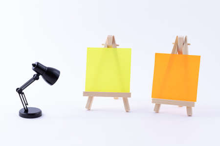 Two Wooden Easels Miniature with Blank Colored Square Canvas or Memo Paper - Mockup. Mini Wooden Stand with Clean Artboard and Small Black Table Lamp on White Background, Copy Spaceの写真素材