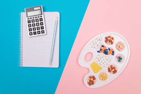 Global Pharmaceutical Industry and Treatment Costs - Different Colored Pills, Tablets and Capsules on White Art Palette with Calculator and Notepad Lying on Split Blue and Pink Background, Flat Layの写真素材