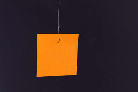Mockup of a Blank Orange Memo Paper with Copy Space Hanging on a Fishing Hook Against the Black Background. Sticky Note Templateの写真素材