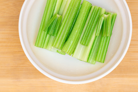Fresh Chopped Celery Sticks with Water Drops in White Bowl on Bamboo Cutting Board - Top View. Vegan and Vegetarian Culture. Raw Food. Healthy Diet with Negative Calorie Contentの写真素材