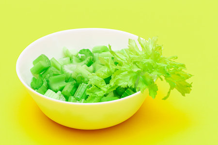 Fresh Chopped Celery Pieces with Water Drops in White Bowl on Yellow Background. Vegan and Vegetarian Culture. Raw Food. Healthy Diet with Negative Calorie Contentの写真素材