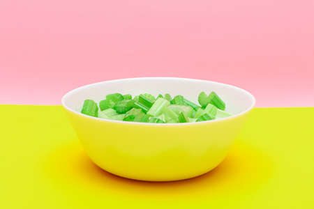 Fresh Chopped Celery Pieces with Water Drops in White Bowl on Yellow Background. Vegan and Vegetarian Culture. Raw Food. Healthy Diet with Negative Calorie Contentの写真素材