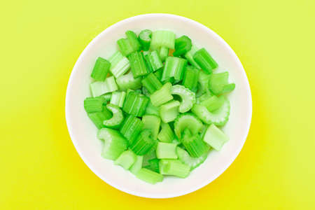 Fresh Chopped Celery Pieces with Water Drops in White Bowl on Yellow Background. Vegan and Vegetarian Culture. Raw Food. Healthy Diet with Negative Calorie Contentの写真素材