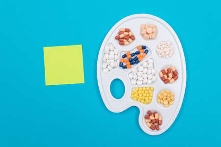 Global Pharmaceutical Industry and Medicinal Products - Different Colored Pills, Tablets and Capsules on White Art Palette with Blank Sticky Note and Copy Space Lying on Blue Background, Flat Layの写真素材