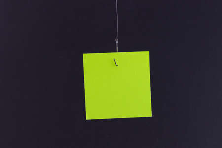 Mockup of a Blank Green Memo Paper with Copy Space Hanging on a Fishing Hook Against the Black Background. Reminder or To Do List. Sticky Note Templateの写真素材