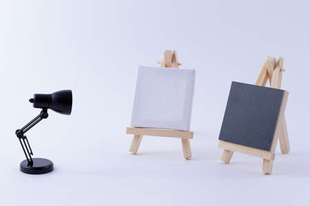 Two Wooden Easels Miniature with Blank Black and White Square Canvas for Artists and Painters - Mockup. Mini Wooden Stand with Clean Artboard and Small Black Table Lamp on White Background, Copy Spaceの写真素材