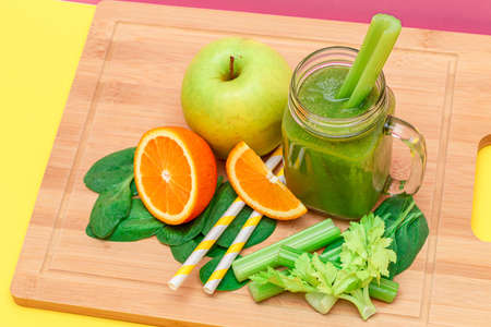 Fresh Green Smoothie of Apple, Celery, Spinach and Orange in Glass Smoothie Jar with Yellow Cocktail Straw on Wooden Cutting Board. Vegan Detox Drink. Vegetarian Culture. Healthy Eating and Fruit Dietの写真素材