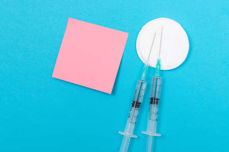 Vaccination, Immunology or Revaccination Concept - Two Medical Disposable Syringe Lying on Blue Table in Doctors Office in a Hospital or Clinic. Blank Pink Sticky Note - Mock Up with Copy Spaceの写真素材