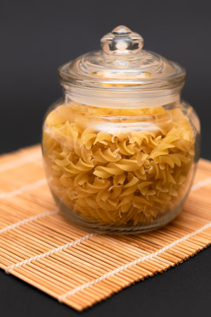 Uncooked Fusilli Pasta in Glass Jar on Black Background. Raw and Dry Macaroni. Unhealthy and Fat Food. Italian Cultureの写真素材