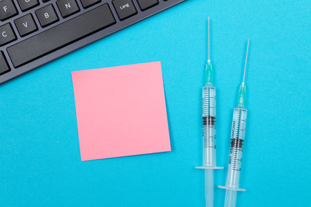 Vaccination, Immunology or Revaccination Concept - Two Medical Disposable Syringe Lying on Blue Table in Doctors Office in a Hospital or Clinic. Blank Pink Sticky Note - Mock Up with Copy Spaceの写真素材