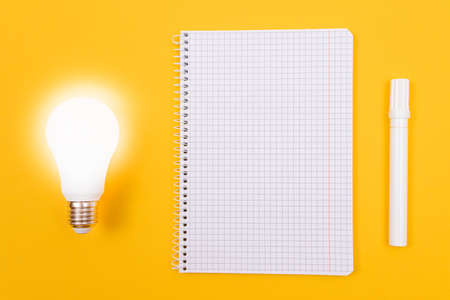 Glowing Power Saving Lamp with Notepad Lying on Yellow Table, Blank Template with Copy Space - Top View, Flat Layの写真素材