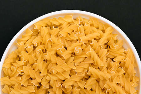 Uncooked Fusilli Pasta Lying on White Plate on Black Background. Raw and Dry Macaroni. Unhealthy and Fat Food - Top Viewの写真素材