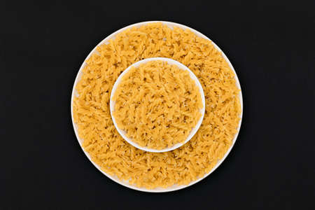 Uncooked Fusilli Pasta Lying on White Plate on Black Background. Raw and Dry Macaroni. Unhealthy and Fat Food - Top Viewの写真素材