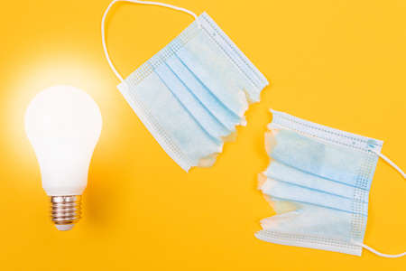 Glowing Power Saving Lamp with Torn Medical Face Mask Lying on Yellow Background - Anti Mask Conceptの写真素材