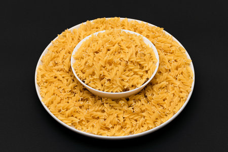Uncooked Fusilli Pasta Lying on White Plate on Black Background. Raw and Dry Macaroni. Unhealthy and Fat Foodの写真素材