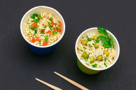 Prepared Instant Noodles with Green Pea, Carrot and Greens in Blue and Green Cups with Chopsticks on Black Background. Quick Lunch or Unhealthy Fast Foodの写真素材