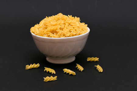 Uncooked Fusilli Pasta in White Bowl on Black Table. Raw and Dry Macaroni. Unhealthy and Fat Food. Italian Cultureの写真素材