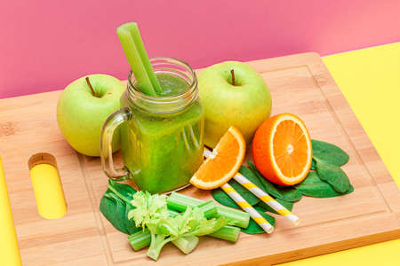 Fresh Green Smoothie of Apple, Celery, Spinach and Orange in Glass Smoothie Jar with Yellow Cocktail Straw on Wooden Cutting Board. Vegan Detox Drink. Vegetarian Culture. Healthy Eating and Fruit Dietの写真素材