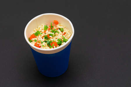 Prepared Instant Noodles with Carrot and Greens in Blue Cup on Black Background. Quick Lunch or Unhealthy Fast Foodの写真素材