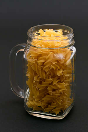 Uncooked Fusilli Pasta in Glass Jar on Black Background. Raw and Dry Macaroni. Unhealthy and Fat Food. Italian Cultureの写真素材