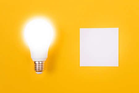 Glowing Power Saving Lamp with White Note Paper Lying on Yellow Table, Blank Template with Copy Space - Top View, Flat Layの写真素材