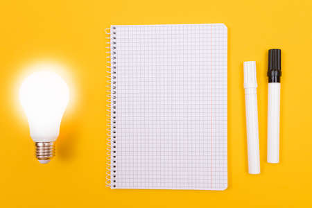 Glowing Power Saving Lamp with Notepad Lying on Yellow Table, Blank Template with Copy Space - Top View, Flat Layの写真素材