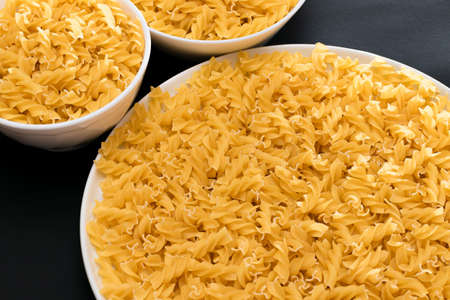 Uncooked Fusilli Pasta Lying on White Plate on Black Background. Raw and Dry Macaroni. Unhealthy and Fat Foodの写真素材