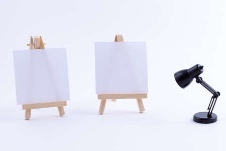 Two Wooden Easels Miniature with Blank White Square Canvas for Artists and Painters - Mockup. Mini Wooden Stands with Clean Artboard and Small Black Table Lamp on White Background, Copy Spaceの写真素材